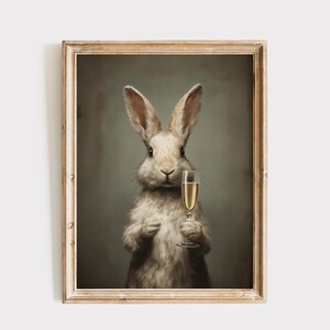 May include: A framed print featuring a rabbit holding a champagne flute. The rabbit has light gray fur and is set against a muted green background. The champagne in the glass is a golden color, and the frame is a light wood tone.