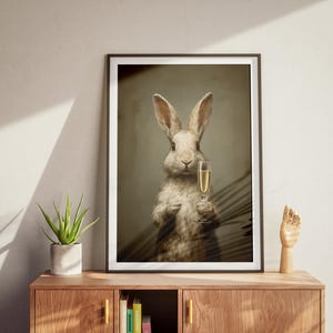 May include: Framed art print featuring a rabbit holding a champagne glass. The rabbit has light brown fur and is set against a muted background. The print is in a black frame and is displayed on a wooden cabinet.