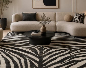 May include: A rectangular area rug featuring a black and white zebra stripe pattern. The rug is positioned in a living space beneath a round black coffee table, a curved beige sofa, and a matching beige armchair, creating a focal point.