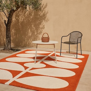 May include: An outdoor scene featuring a vibrant orange and white patterned rug. A small white table with a woven basket and a black chair are on the rug. A tree and a beige wall complete the scene.