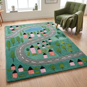 May include: A rectangular area rug featuring a green background with a winding gray road, a blue pond, and various pink houses with black roofs. The design includes scattered green trees and bushes, creating a stylized map-like landscape pattern.