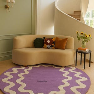 Op de afbeelding: Een woonkamer met een gebogen beige bank en sierkussens, een rond paars tapijt met een golvende crèmekleurige rand en een kleine zwarte bijzettafel. Het tapijt heeft de tekst "The loom lane". Een vaas met zonnebloemen staat op de tafel.