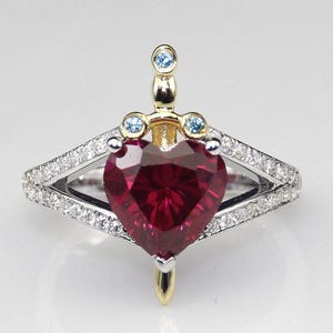 Può includere: Anello a forma di cuore con rubino e design a spada. Il rubino è il punto focale, incastonato in una fascia d'oro e argento. La spada presenta pietre blu e un'elsa dorata, aggiungendo un tocco unico al gioiello.