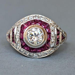 Peut inclure: Une bague de style vintage avec une pierre centrale ronde et transparente, entourée de pierres rouges profondes taillées en carré. La bague a un anneau argenté avec de petites pierres d'accent claires. Le design est symétrique et orné, avec un attrait classique et élégant.
