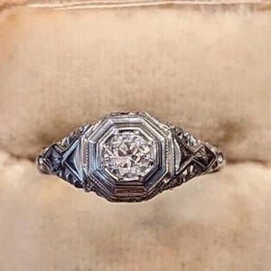 May include: An antique silver ring with a central, round-cut diamond. The ring features an octagonal setting with intricate filigree detailing on the band. The ring is displayed inside a red velvet jewelry box.