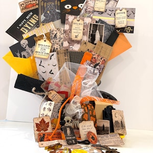 May include: A Halloween-themed craft bouquet featuring assorted paper cards, tags, and ephemera with motifs of skeletons, pumpkins, bats, and black cats. The arrangement includes orange and black ribbons, tulle, and various small buttons and charms.