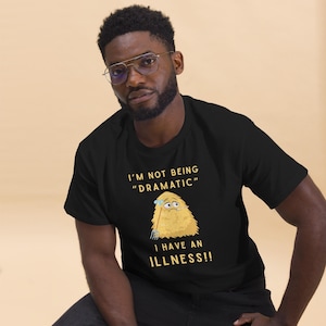 Peut inclure: T-shirt noir avec le texte "I'M NOT BEING 'DRAMATIC' I HAVE AN ILLNESS!!" et une illustration de dessin animé d'une créature jaune et poilue avec une expression inquiète. Le t-shirt est fait d'une matière douce.