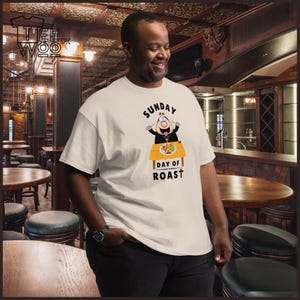 Puede incluir: Una camiseta color crema con un sacerdote de dibujos animados sosteniendo cuchillos sobre una mesa con comida. El texto "SUNDAY DAY OF ROAST" está impreso en negro y naranja. La camiseta la lleva una persona en un restaurante.