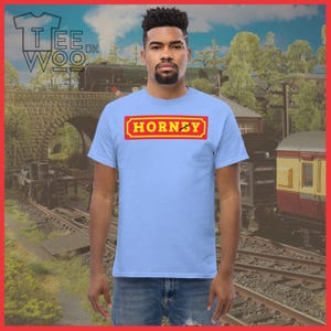 May include: A light blue t-shirt with the word "HORNBY" in a red and yellow rectangular design. The shirt is being worn by a person. The background features a train and railway tracks.