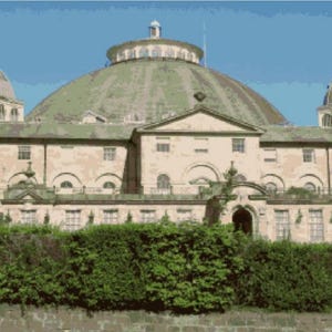 May include: A large, historic building with a prominent green dome and two smaller domes. The building has a light-colored facade with multiple windows and is surrounded by greenery under a clear blue sky.