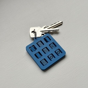 May include: A blue square keyring with nine recessed designs, alongside two silver keys. The keyring is made of a solid material and has a textured surface. The keys are attached to the keyring.
