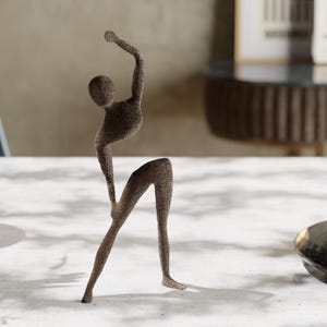 Könnte beinhalten: Eine braune, abstrakte Skulptur einer Person in einer Tanzpose. Die Figur hat einen Arm erhoben und ein Bein ausgestreckt, mit einer strukturierten Oberfläche. Die Skulptur steht auf einer weißen Oberfläche.