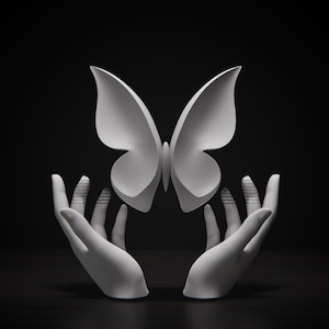 Puede incluir: Una escultura blanca de una mariposa con las alas extendidas, aparentemente sostenida por dos manos. Las manos también son blancas y están posicionadas para sostener la mariposa. El fondo es negro liso.