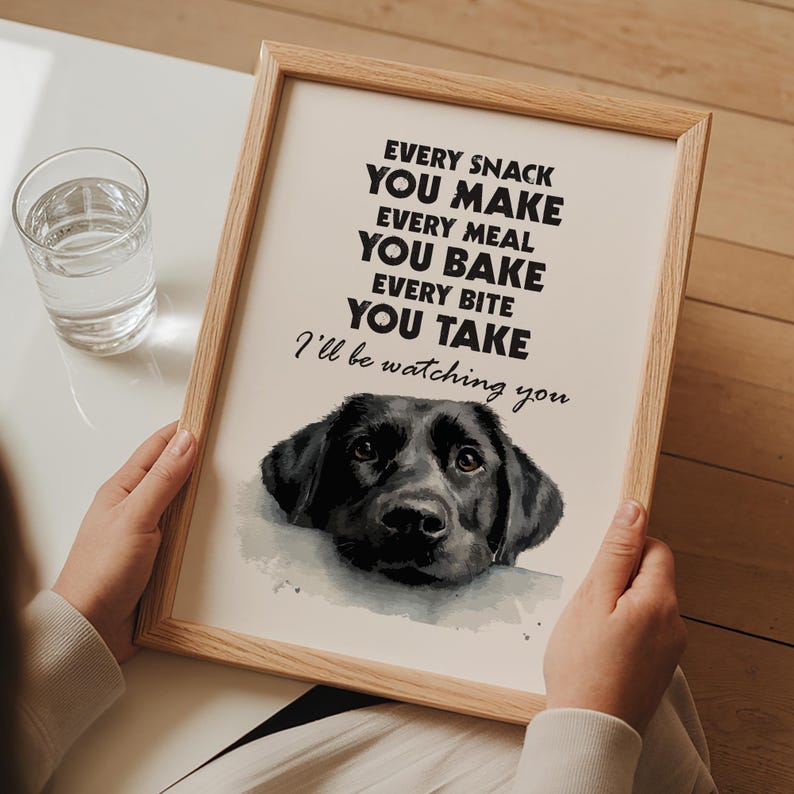 May include: A framed print with a light wood frame. The print features the text "EVERY SNACK YOU MAKE, EVERY MEAL YOU BAKE, EVERY BITE YOU TAKE, I'll be watching you" above a watercolor image of a black Labrador Retriever.
