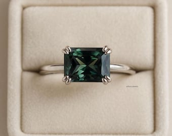 May include: A silver ring featuring a rectangular, emerald-cut, dark green gemstone. The gemstone is held in place by four prongs. The ring is presented in a beige ring box. The brand name "wilsonjewels" is visible on the right side.