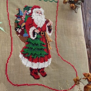 May include: Needlepoint Christmas stocking featuring Santa Claus carrying a sack of toys and a Christmas tree. The design is stitched on a beige canvas with a red outline. Santa is dressed in a red suit with white trim and boots.