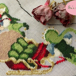 May include: A cross-stitch embroidery of two green and red figures holding a brown sack, with a red and gold sleigh in the foreground. The embroidery is on a white canvas with a pink label that reads "Su's Treasure".