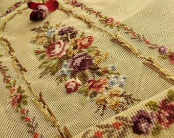 May include: A beige canvas needlepoint with a floral design. The design features a large bouquet of pink, purple, and yellow flowers with green leaves. The flowers are arranged in a symmetrical pattern around a central archway. The canvas is surrounded by a border of smaller flowers and leaves.