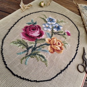 Puede incluir: Un lienzo beige con un marco ovalado negro, que presenta un bordado de punto de cruz de un ramo de flores. El ramo incluye una rosa rosa, una rosa amarilla y flores azules.