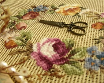 Pode incluir: Um close-up de uma tela bege com um desenho de bordado floral. O bordado apresenta flores rosa, roxas, amarelas, azuis e verdes. Um par de tesouras de prata antigas está sobre a tela.