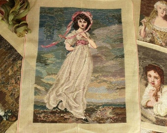 May include: A needlepoint canvas featuring a woman in a white dress with a pink sash and a pink hat. The woman is standing in a landscape with a blue sky and clouds. The canvas is surrounded by a beige frame.