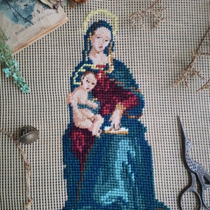Könnte beinhalten: Eine Nadelmalerei, die die Jungfrau Maria zeigt, die das Jesuskind hält. Die Stickerei ist in einem hellbraunen Holzrahmen gerahmt und befindet sich auf einem beigefarbenen Leinwandhintergrund.
