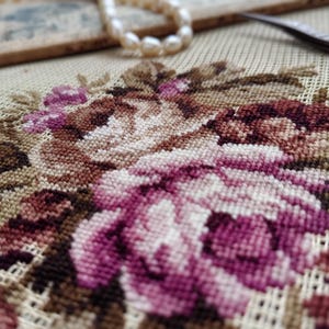May include: Close-up of a floral embroidery pattern on a beige fabric. The design features pink and brown roses with intricate details.