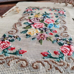 Puede incluir: Un lienzo beige con un diseño de bordado floral. El bordado presenta una variedad de flores en tonos de rosa, rojo, amarillo, azul y verde. Las flores están dispuestas en un patrón simétrico y están rodeadas por un borde marrón.