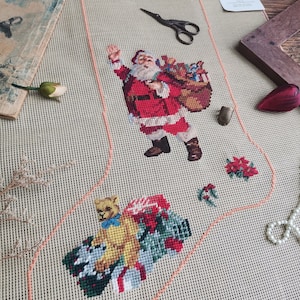 May include: A beige canvas with a red and white Santa Claus design, a teddy bear, and a bag of presents. The canvas is outlined in pink and is ready for needlepoint.