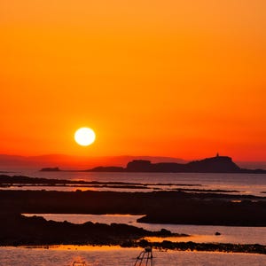 以下が含まれることがあります： 鮮やかな夕焼けの海、明るいオレンジ色の空と大きな太陽。手前にボートがあり、遠くには陸地のシルエットが見えます。水面は夕焼けの暖色を映し出しています。