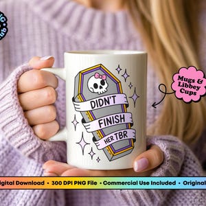 May include: White ceramic mug featuring a coffin design with a skull, a pink bow, and the text "DIDN'T FINISH HER TBR." The mug has a handle and is decorated with stars. The mug is held by a person.