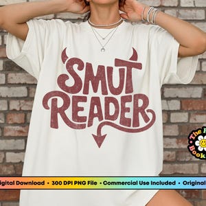 May include: Cream-colored t-shirt with the text "SMUT READER" in a distressed, maroon font. The design includes devil horns and a downward-pointing arrow. A small logo is visible on the bottom right.