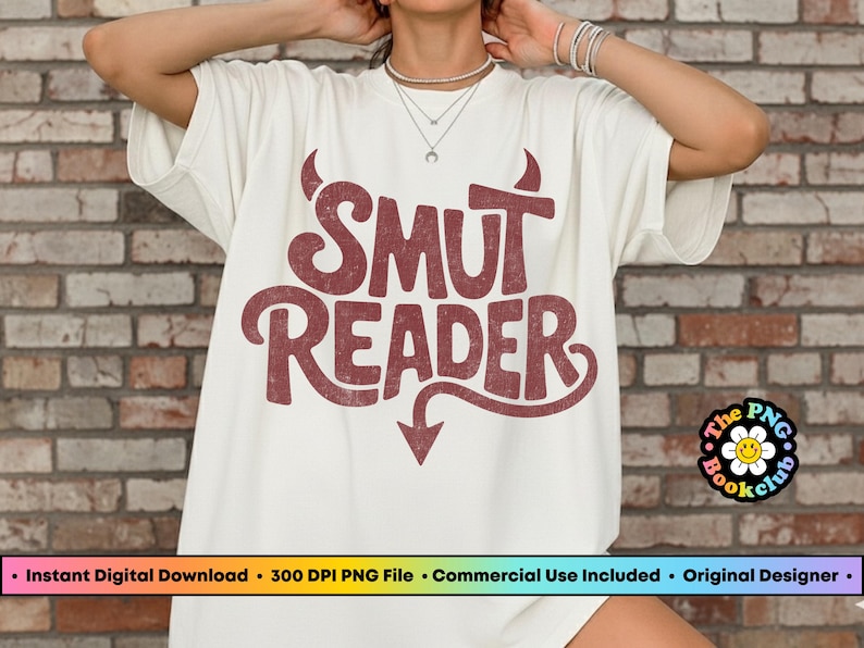 May include: Cream-colored t-shirt with the text "SMUT READER" in a distressed, maroon font. The design includes devil horns and a downward-pointing arrow. A small logo is visible on the bottom right.