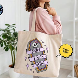 May include: Beige tote bag with a coffin graphic, a skull with a pink bow, and the text "Didn't Finish Her TBR". The tote bag has a light purple ribbon design and star accents. The tote bag is a book club item.