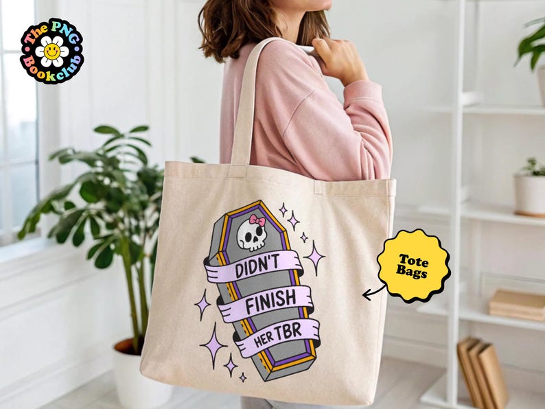 May include: Beige tote bag with a coffin graphic, a skull with a pink bow, and the text "Didn't Finish Her TBR". The tote bag has a light purple ribbon design and star accents. The tote bag is a book club item.