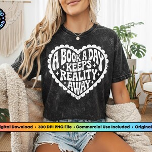 Pode incluir: Camiseta preta com um gr&aacute;fico branco em forma de cora&ccedil;&atilde;o que diz "A BOOK A DAY KEEPS REALITY AWAY". A camiseta tamb&eacute;m tem o texto "The PNG Bookclub" em um logotipo circular. A camiseta &eacute; usada por uma pessoa.