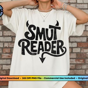 May include: Off-white t-shirt with the text "SMUT READER" in a distressed black font. The design includes devil horns above the word "SMUT" and an arrow pointing down below "READER". The shirt is a casual style.