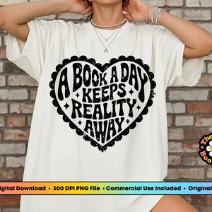 Pode incluir: Camiseta branca com um desenho de cora&ccedil;&atilde;o preto que diz "A BOOK A DAY KEEPS REALITY AWAY" em uma fonte retr&ocirc;. A camiseta tem gola redonda e mangas curtas. O logotipo PNG Bookclub &eacute; vis&iacute;vel.