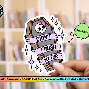 May include: A sticker featuring a coffin design with a skull wearing a pink bow. The coffin has a banner that reads "DIDN'T FINISH HER TBR". The sticker is held by a hand. The background is a bookshelf.