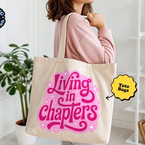 May include: A beige canvas tote bag featuring a vibrant pink graphic design with the text "Living in chapters" in a stylized, bold script font, accented by small white star shapes, with two sturdy shoulder straps for carrying items.