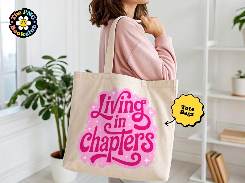 May include: A beige canvas tote bag featuring a vibrant pink graphic design with the text "Living in chapters" in a stylized, bold script font, accented by small white star shapes, with two sturdy shoulder straps for carrying items.