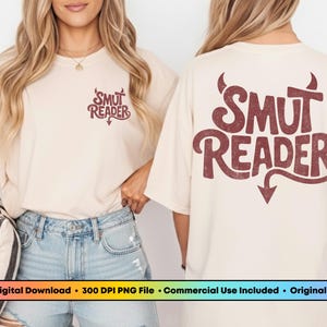 May include: Cream-colored t-shirt with the text "SMUT READER" in a retro font, featuring a devil's tail and horns design. The shirt is worn with denim shorts.