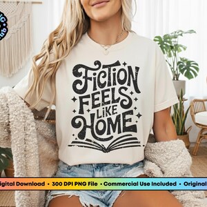 May include: Cream-colored t-shirt with the text "Fiction Feels Like Home" in a black, distressed font. The design includes stars and an open book. The shirt is part of a book club collection.