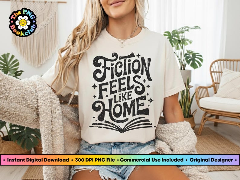 May include: Cream-colored t-shirt with the text "Fiction Feels Like Home" in a black, distressed font. The design includes stars and an open book. The shirt is part of a book club collection.