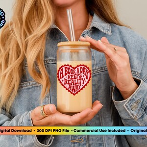 Pode incluir: Um copo de vidro transparente com tampa de bambu e canudo de vidro, cheio de uma bebida cremosa. O copo apresenta um design em forma de cora&ccedil;&atilde;o vermelho com o texto "A Book A Day Keeps Reality Away". A imagem &eacute; contra uma jaqueta jeans.