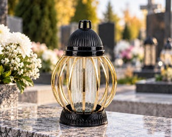 Könnte beinhalten: Eine Grablampe mit einem klaren, vertikal gerippten Glaszylinder, der von einem goldfarbenen Metallkäfig umgeben ist. Die Lampe verfügt über einen schwarzen Kunststoffsockel und einen passenden schwarzen, gewölbten Deckel mit Griff.