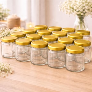 Op de afbeelding: Een verzameling heldere glazen potten met goudkleurige deksels. De potten zijn gerangschikt op een lichtgekleurd houten oppervlak, met een zachte achtergrond van bloemen en kaarsen. De potten zijn leeg en klaar om te worden gevuld.