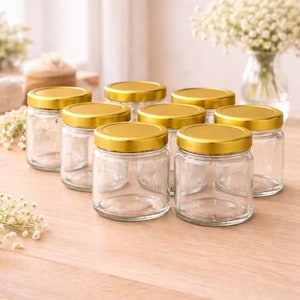 Op de afbeelding: Zeven heldere glazen potten met gouden deksels staan op een licht houten oppervlak. De achtergrond bevat bloemen en decoratieve items. De potten zijn leeg en klaar voor gebruik. Elke pot is ongeveer 8 cm hoog.