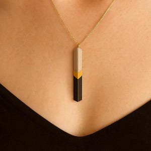 May include: A gold chain necklace with a geometric pendant. The pendant is a vertical rectangle with sections of black, gold, and grey. The necklace is worn against a black top.