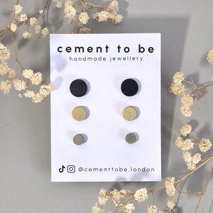 May include: A white card with the text "cement to be handmade jewellery" displays six round stud earrings in black, gold, and grey. The earrings are arranged in pairs. The card also includes social media handles. Dried flowers are in the background.